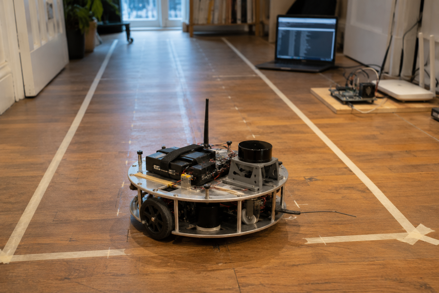 Alpha mid-lane on the hall boards, taped lane edges and calibration marks running away toward the door, the trace laptop glowing out of focus at the back, the dock rig and access point on their plywood board at the skirting.